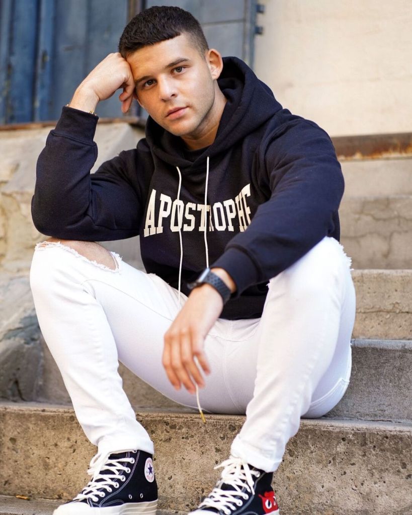 Josh Riquelme sits on concrete stairs in SoHo, NYC, gazing at you. Josh wears a navy blue hoodie with Apostrophe in white letters on the front and white, distressed jeans.